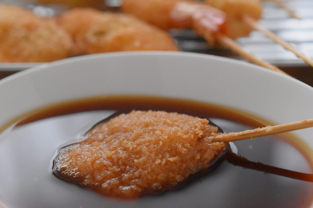 串カツソース（ウスターソースを水で溶いたソース）をつけて串カツを食べる写真-Photo(C)Kumitatetsushin
